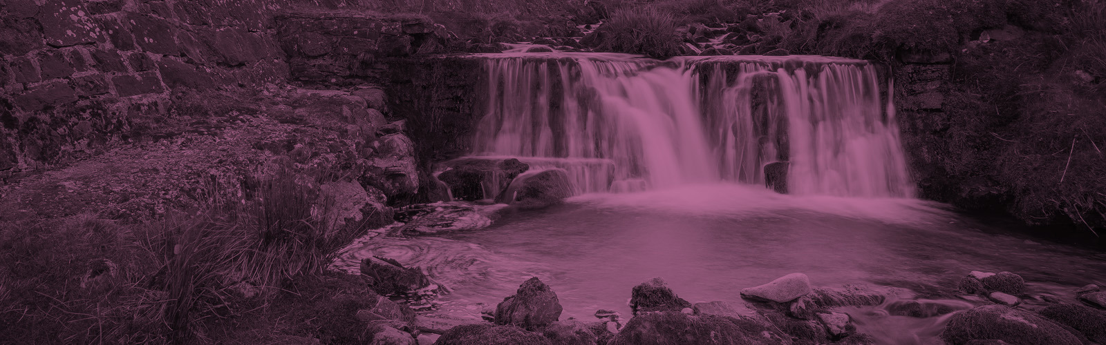 Waterfall cascading over rocks into  pool, surrounded by green hills and mountains.